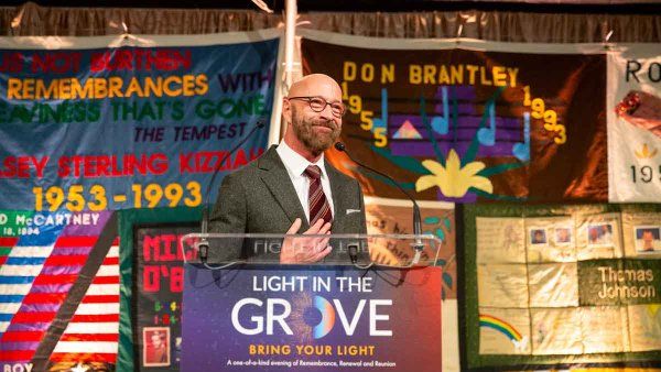 Dan Bernal smiles as he stands at a podium that has a sign that reads "Light in the Grove." In the background are multicolored flags with names of previous award winners.