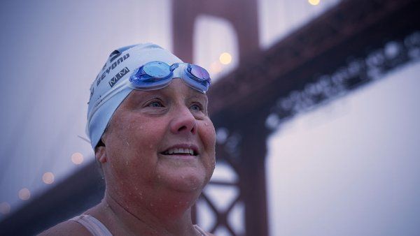 Amy Appelhans Gubser wears a swim cap and goggles and stands in front of the Golden Gate Bridge at sunrise.