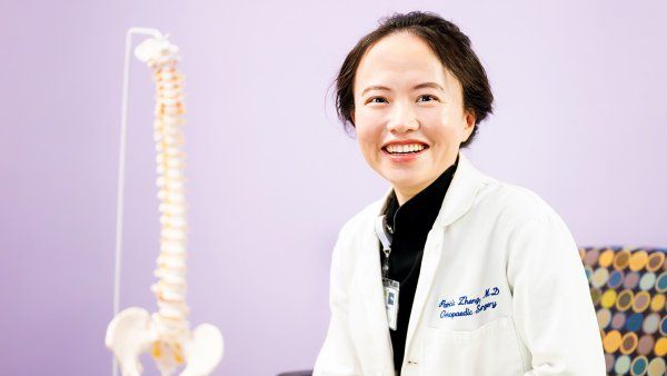Dr. Zheng, smiling in a white lab coat, seated beside a spine model