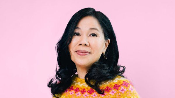 Joanne Chun stands in front of a pink background in a colorful, pink and yellow, hand-knitted, patterned sweater.