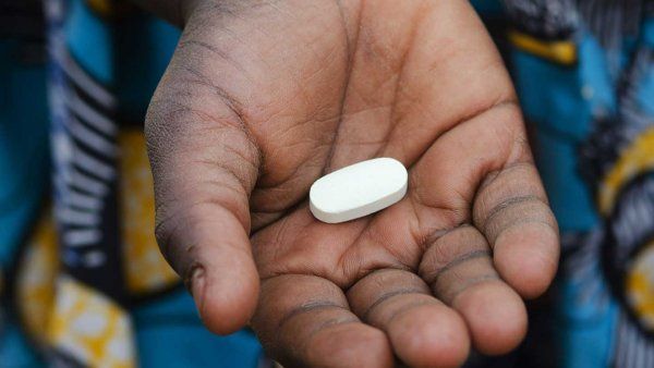 A close-up of a child's hand holding a single, oval-shaped white pill. The child is wearing a brightly patterned blue and yellow fabric.