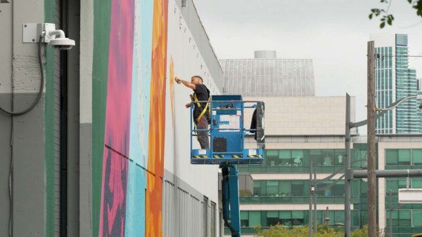 Muralist paints multi-color art on building with city skyline in background.