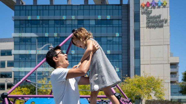 A father smiles as he lifts his daughter at a playground in front of U C S F Benioff Children's Hospital, San Francisco.