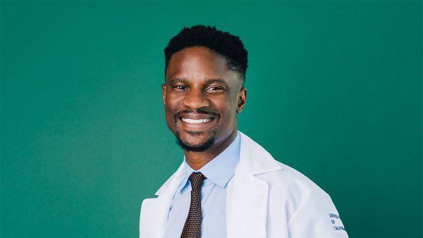 Akinyemi Oni-Orisan wearing a white lab coat and a brown tie.