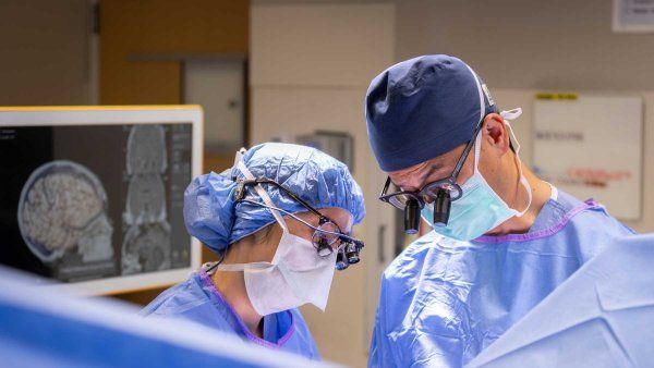 Two neurosurgeons perform surgery with a screen behind them showing a scan of a brain.