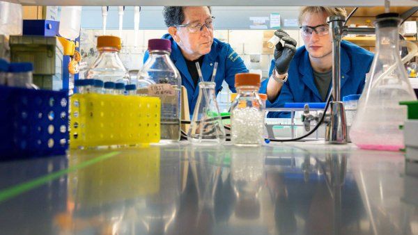 Kevan Shokat and graduate student Hunter Carrell wear blue lab coats and protective goggles as they work at a lab bench.