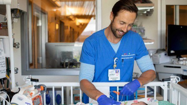 A registered nurse smiles as he tends to a baby in a pediatric intensive care room.