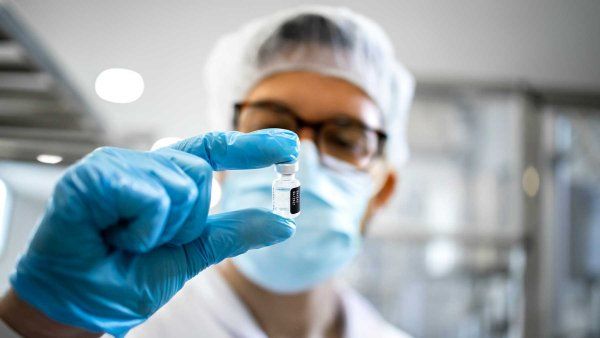 Scientist wearing protective gloves, a mask, and a hair covering examines a small vial in a laboratory.