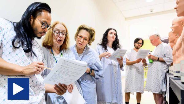 Video play icon: A group of people in hospital gowns stands in a clinical setting, reading and discussing documents while surrounded by medical mannequins.
