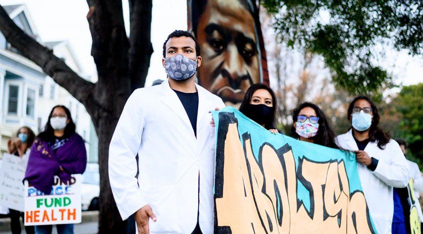 UCSF Nurses gather in Oakland for a Black Health Matters protest; a male Black nurse in a white coat and face mask stands with female nurses, holding a sign that reads “Abolish” in graffiti style; a large mural of George Floyd’s portrait is in the background.