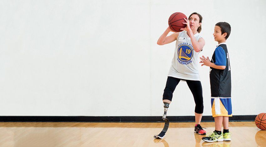 Debrah Bevilacqua (left), has a prosthetic leg from the knee down and wears a Golden State Warriors shirt, shows a young child (right) how to play basketball indoors.
