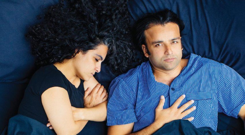 Photo of a man and woman in bed; the woman is fast asleep and the man eats popcorn, wide awake.