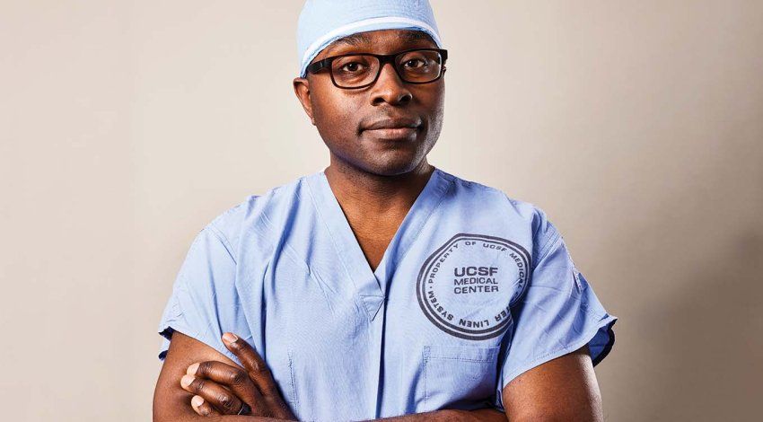 Portrait of neurosurgeon Shawn Hervey-Jumper, standing with his arms crossed in blue surgical scrubs.