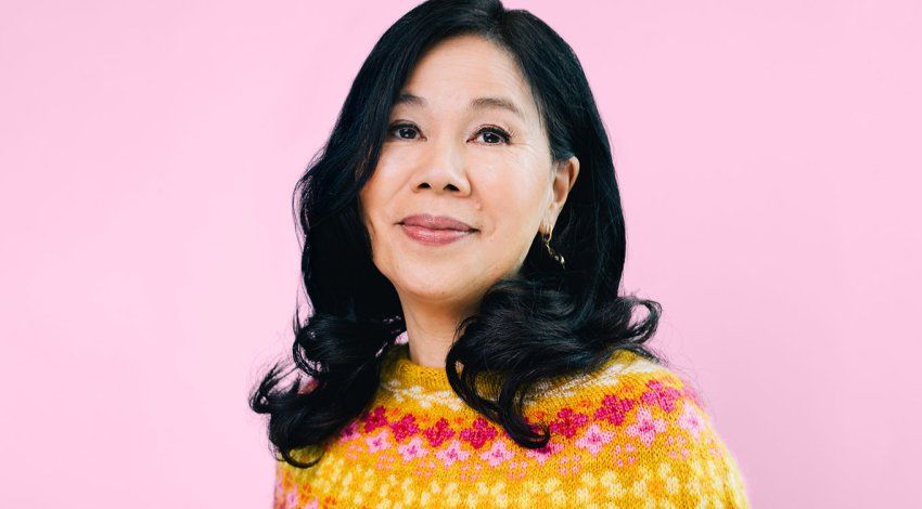Joanne Chun stands in front of a pink background in a colorful, pink and yellow, hand-knitted, patterned sweater.