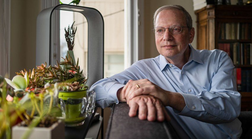 Robert Wachter sits on his couch in his home, resting is arm on the back with succulent pants in the foreground by the window.