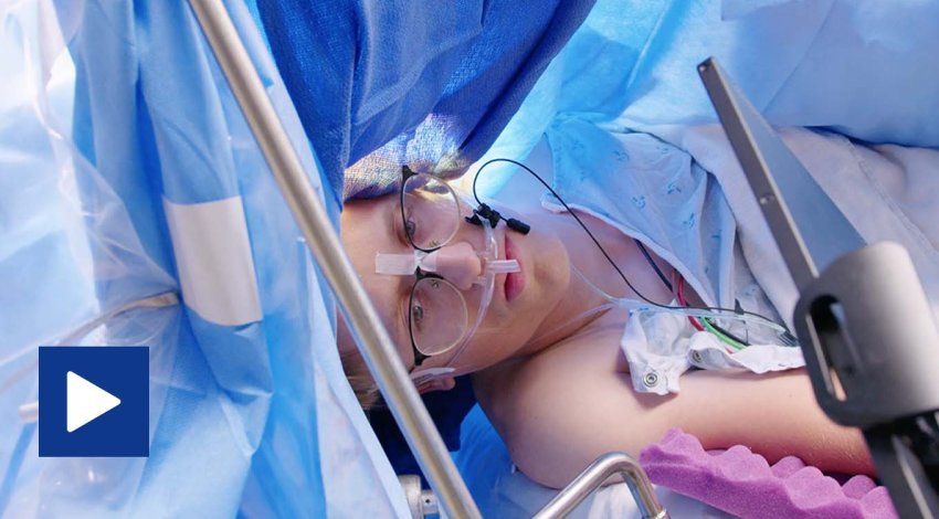 A young man in glasses has his eyes open during surgery as he looks at a screen. Blue surgery drapes are all around him.