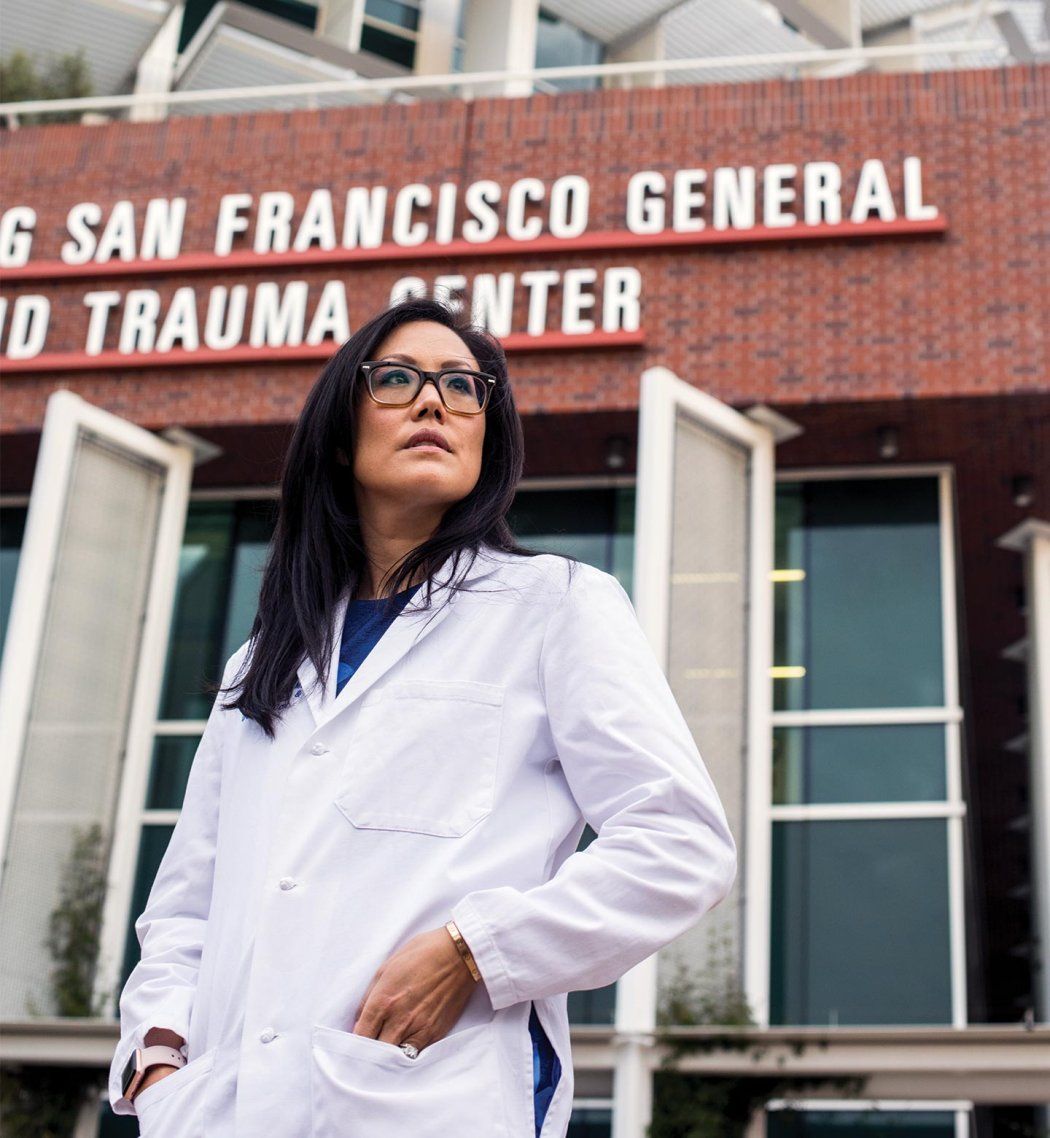 Debbie Yi Madhok wears her doctor’s white coat and stands in front of Zuckerberg San Francisco General Hospital and Trauma Center.