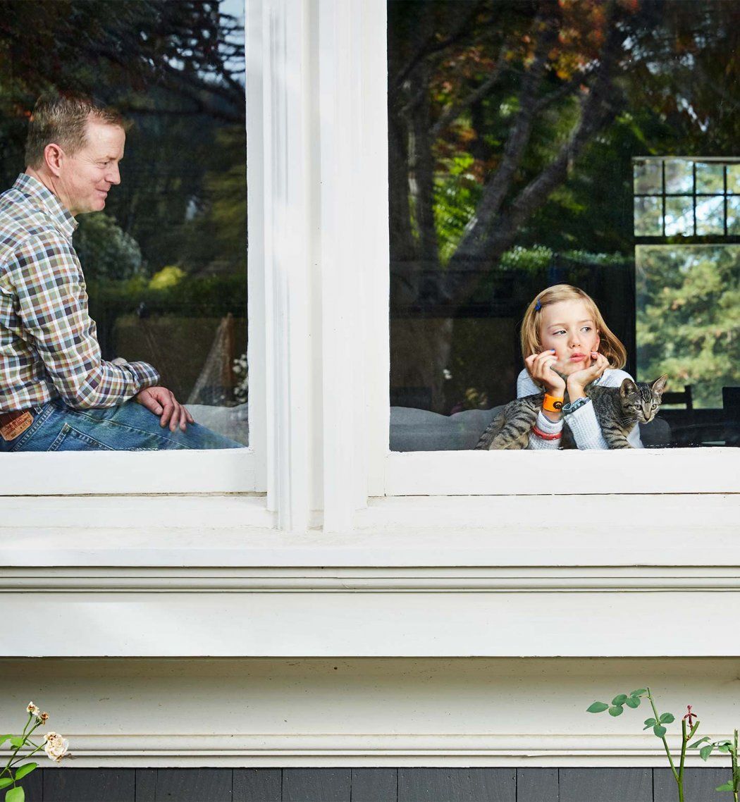 Tim Wood looks down at his daughter, Piper; Piper is holding a cat and looking out the window.