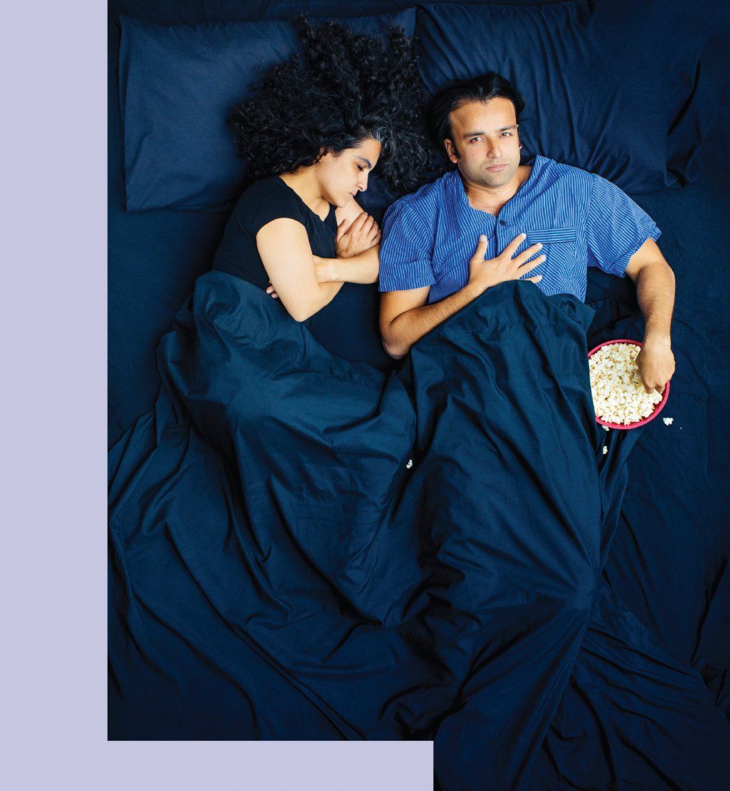 Photo of a man and woman in bed; the woman is fast asleep and the man eats popcorn, wide awake.