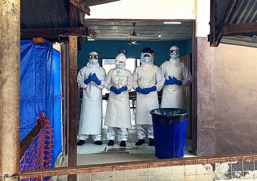 Medical workers in head-to-toe protective suits stand in a doorway of a run-down building with an orange makeshift fence and blue tarp hanging.