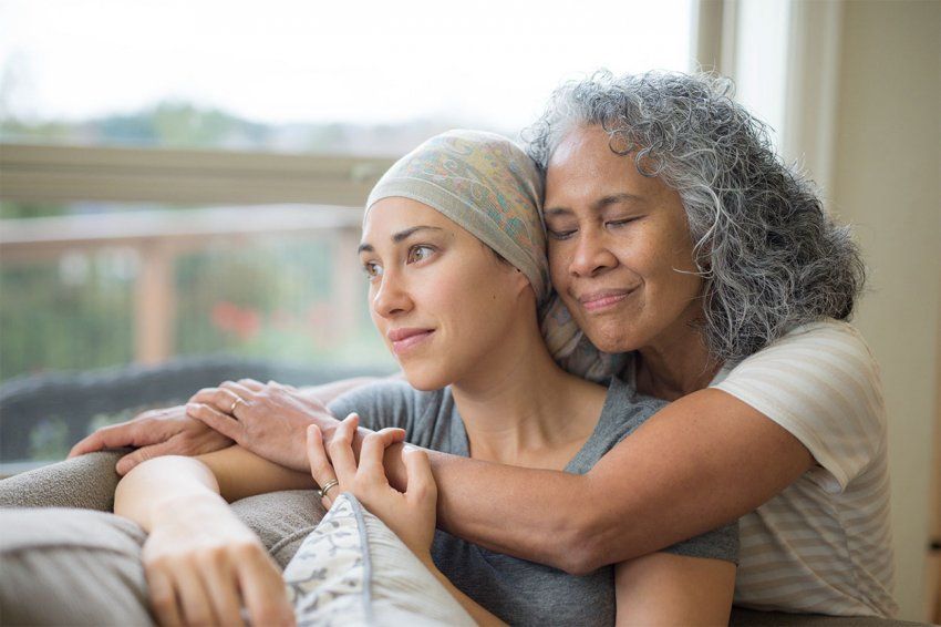 Hawaiian woman in 50s embracing her mid-20s daughter on couch who is fighting cancer.