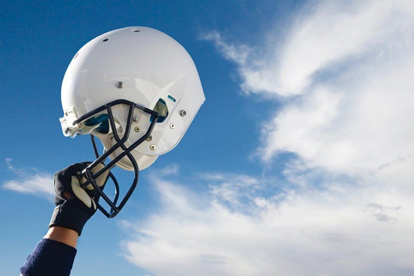 A hand holds up a football helmet in front of a blue sky with clouds.