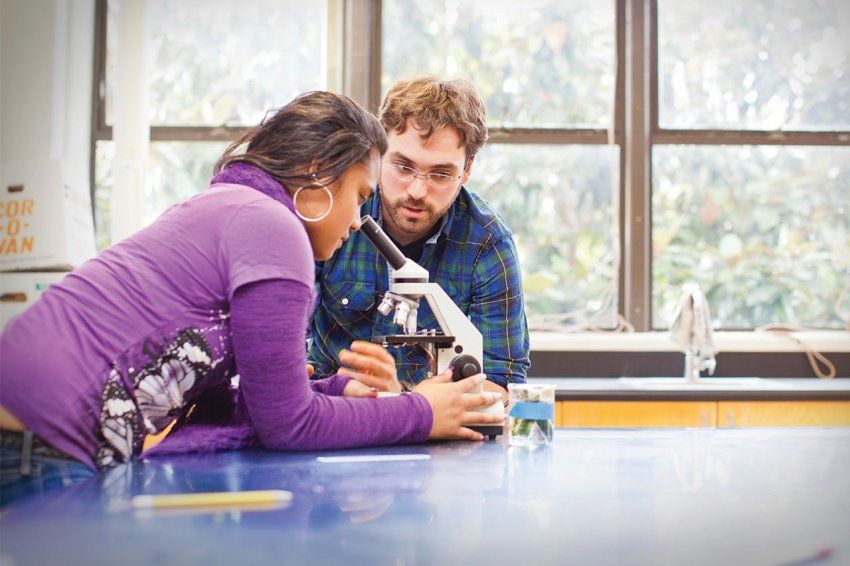 A scientist helps a high school student with a microscope.