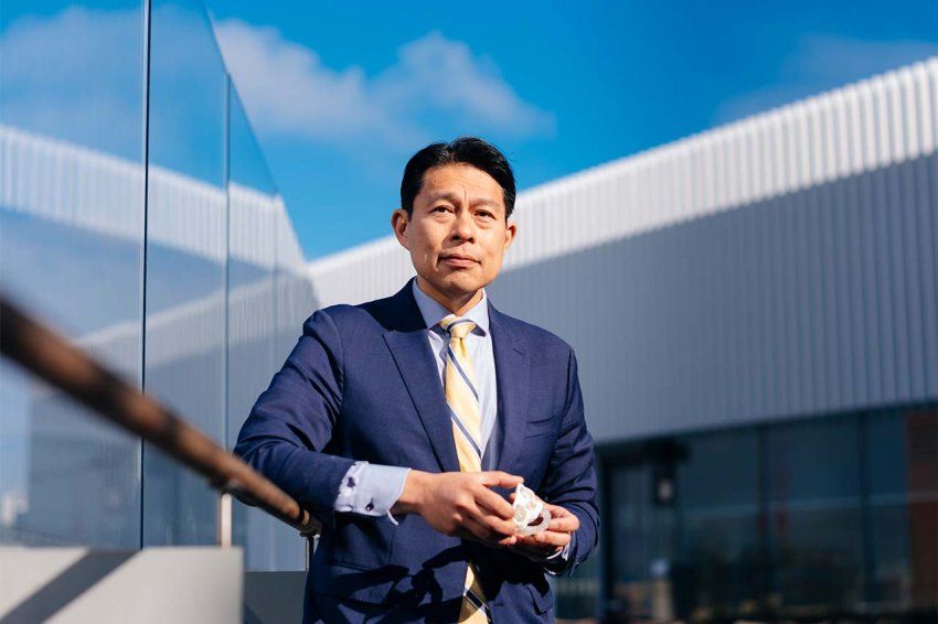 Dr. Lin holds a 3D model of a skull and stands in front of a blue sky and angular buildings.